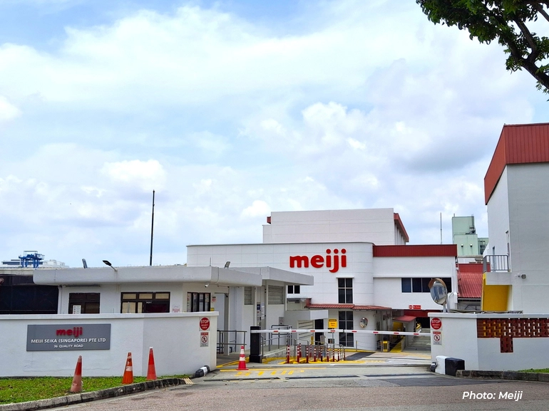 Exterior view of a Meiji factory facility with the company logo on the building, showing the entrance, security gate, and surrounding industrial structures.
