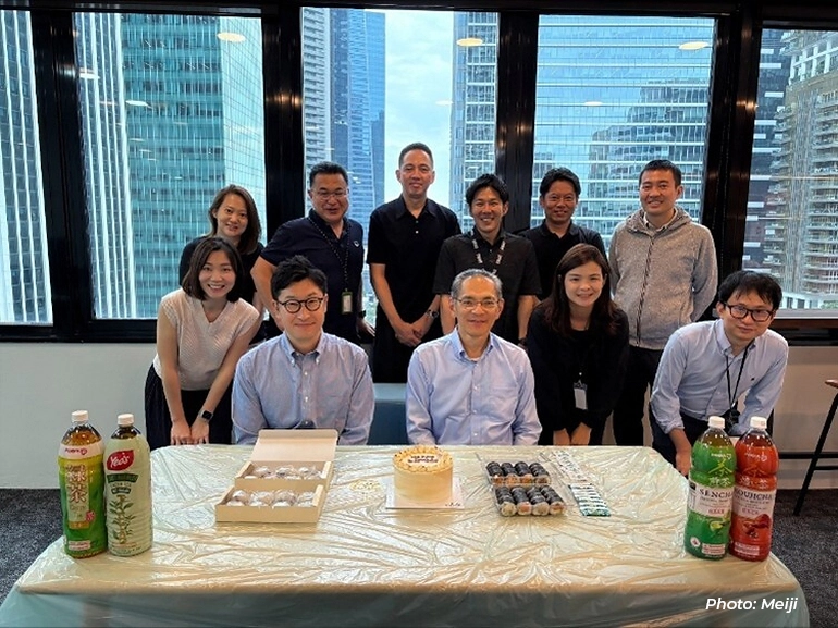 Meiji team members gathered around a table with food and drinks, posing for a group photo in an office setting with city buildings in the background.
