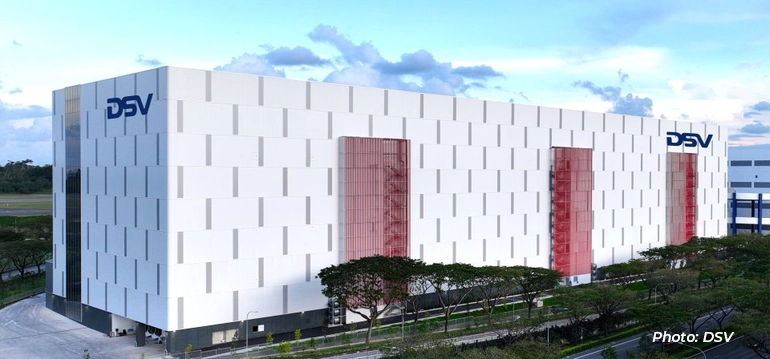 Exterior view of a large DSV logistics facility with a modern white façade and red vertical accents, set beside a roadway with greenery.