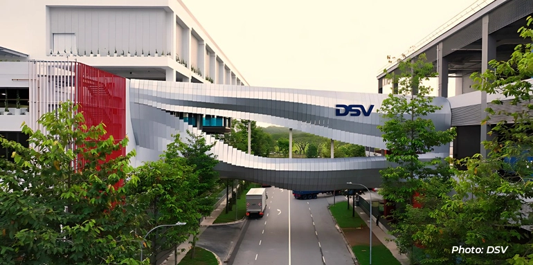 Modern DSV facility with a curved skybridge connecting buildings above a roadway, surrounded by greenery and landscaped grounds.