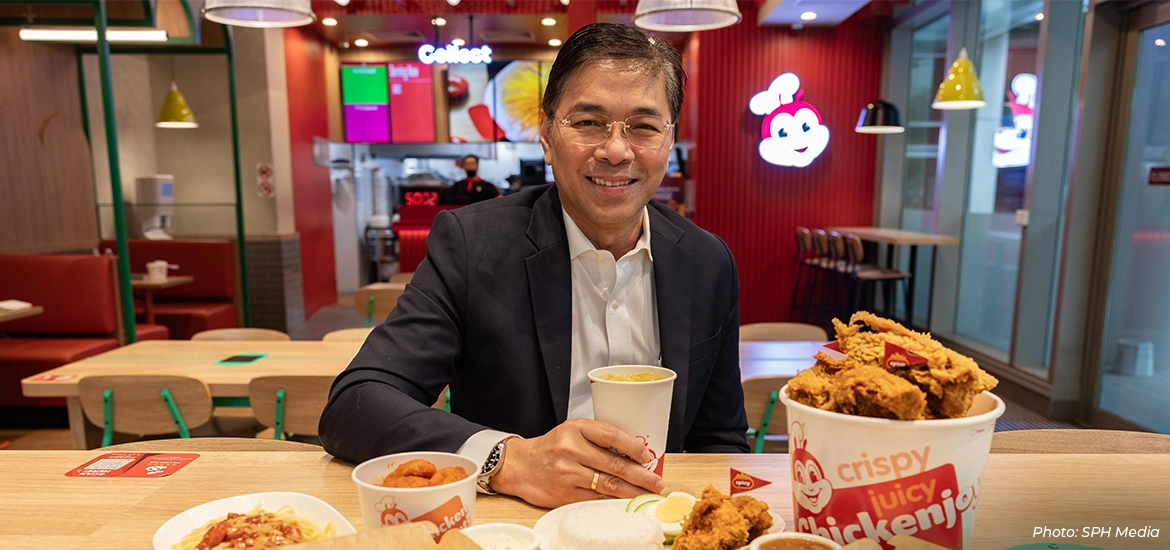 Man in a business suit smiling while dining at a Jollibee restaurant, holding a drink and seated at a table with fried chicken, sides, and branded food packaging, showcasing Singapore’s vibrant food and dining scene.