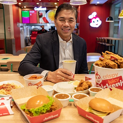 Man in a business suit smiling while dining at a Jollibee restaurant, holding a drink and seated at a table with fried chicken, sides, and branded food packaging, showcasing Singapore’s vibrant food and dining scene.