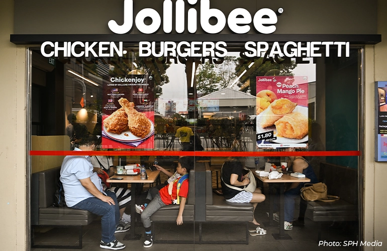 Exterior view of a Jollibee restaurant in Singapore, showing the storefront signage for chicken, burgers, and spaghetti, with diners seated inside behind glass windows and promotional posters for fried chicken and Peach Mango Pie displayed.