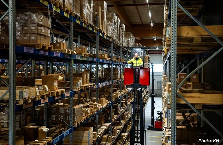 Worker operating a lift in a warehouse aisle with tall shelves stacked with boxes.