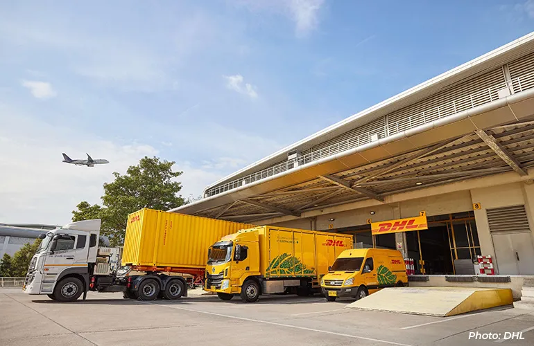 DHL delivery trucks and vans parked at a logistics facility loading bay, with an airplane flying overhead.