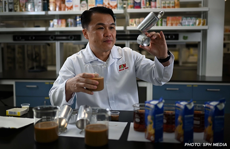 Food technologist in an “F&N Group R&D” lab coat testing beverages in a product‑development lab, holding a stainless measuring cup over a glass of brown drink; sample cups, canisters, and packaged cartons sit on the table with shelves of products behind.