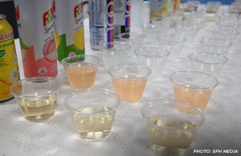 Rows of small plastic cups filled with different flavored beverages for product testing, with F&N and other drink cans in the background.
