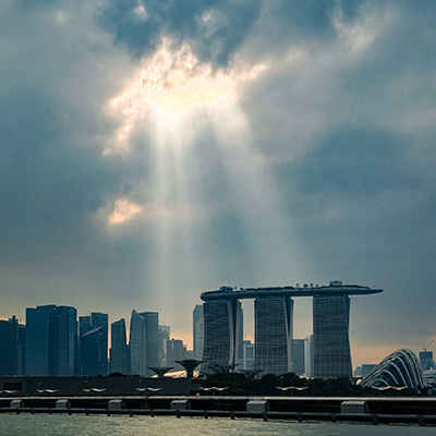Singapore’s Marina Bay skyline at dusk, featuring Marina Bay Sands and surrounding business district towers illuminated by dramatic rays of sunlight breaking through storm clouds.