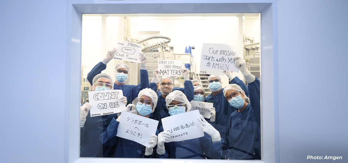 Team members in protective cleanroom attire and face masks posing inside a biopharmaceutical manufacturing facility, holding signs expressing their commitment to patients, innovation, and the company's mission.