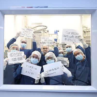 Team members in protective cleanroom attire and face masks posing inside a biopharmaceutical manufacturing facility, holding signs expressing their commitment to patients, innovation, and the company's mission.
