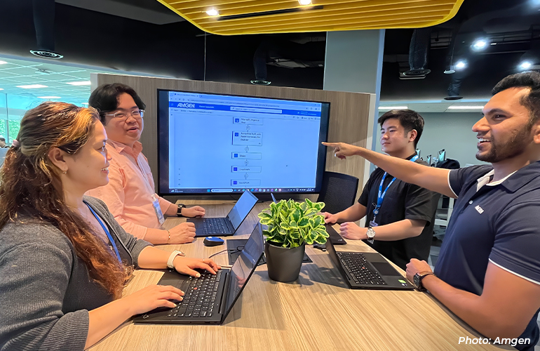 Four employees collaborating around a shared workstation, discussing a digital workflow displayed on a large screen while working on laptops in a modern office environment.