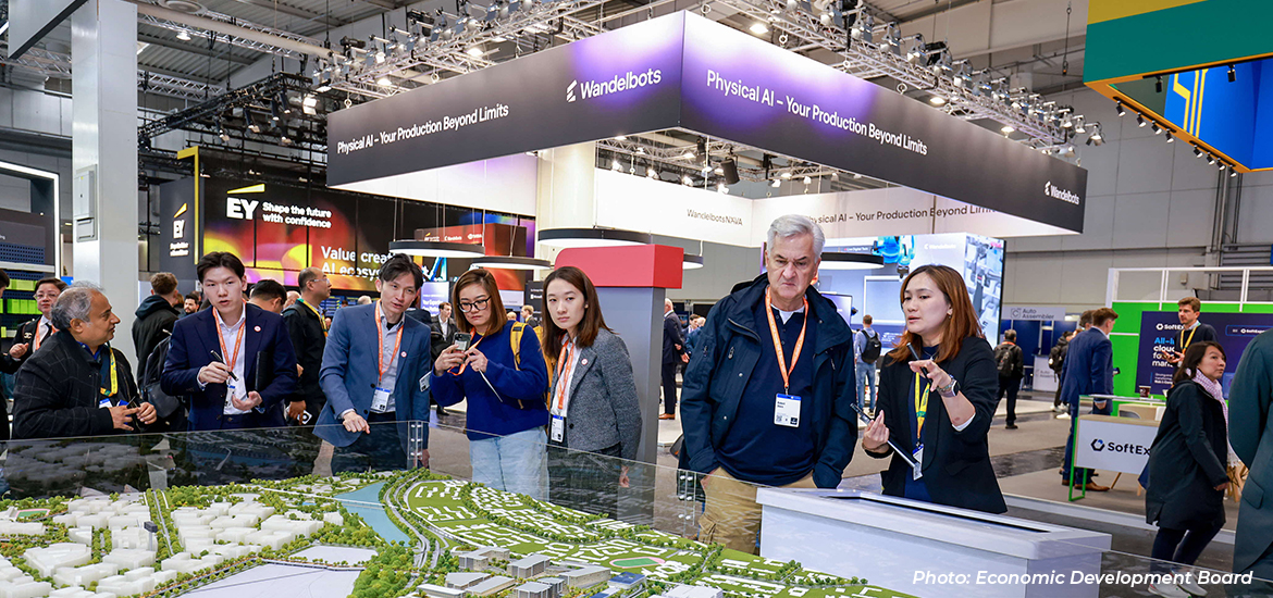 Visitors and exhibitors gathered around a large interactive city model at a technology and innovation exhibition, with booths showcasing AI and advanced manufacturing solutions in the background.