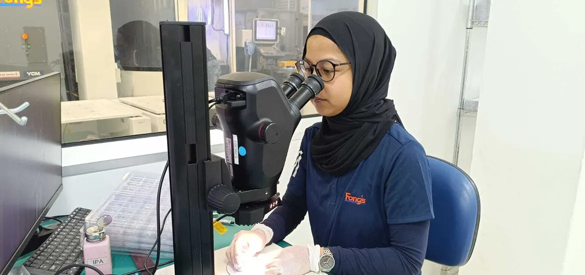 A lab technician in a hijab and gloves uses a microscope to examine small components in a laboratory.