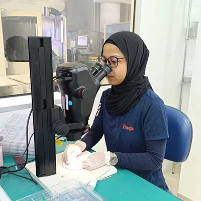 A lab technician in a hijab and gloves uses a microscope to examine small components in a laboratory.