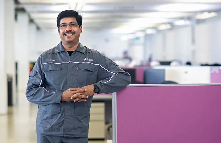 Man wearing a gray Evonik uniform smiling and standing in a brightly lit office with purple partitions.