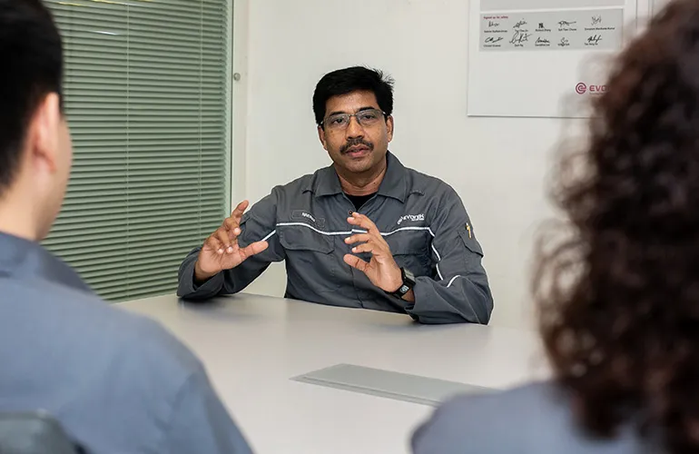 Man in a gray Evonik uniform speaking and gesturing during a meeting with colleagues in a conference room.