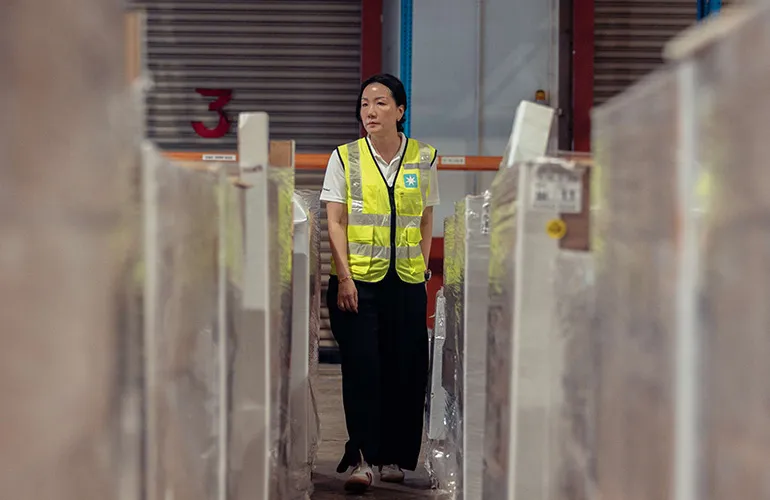 A woman wearing a reflective safety vest walks between stacks of packaged goods in a warehouse.
