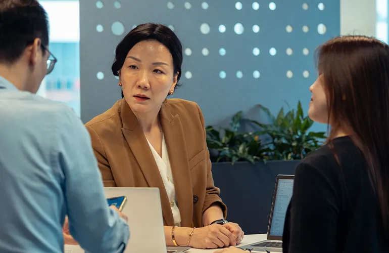 Three colleagues have a discussion around a table with laptops in a modern office setting.