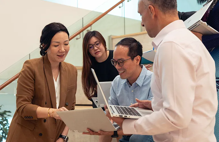 A group of colleagues sit on stairs smiling and collaborating with laptops in hand during an informal meeting.