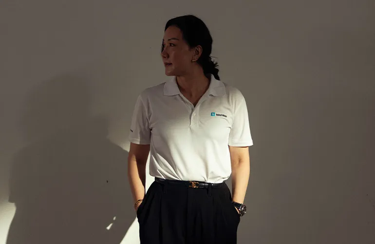A woman wearing a white Maersk polo shirt stands against a plain wall with her hands in her pockets, looking to the side.