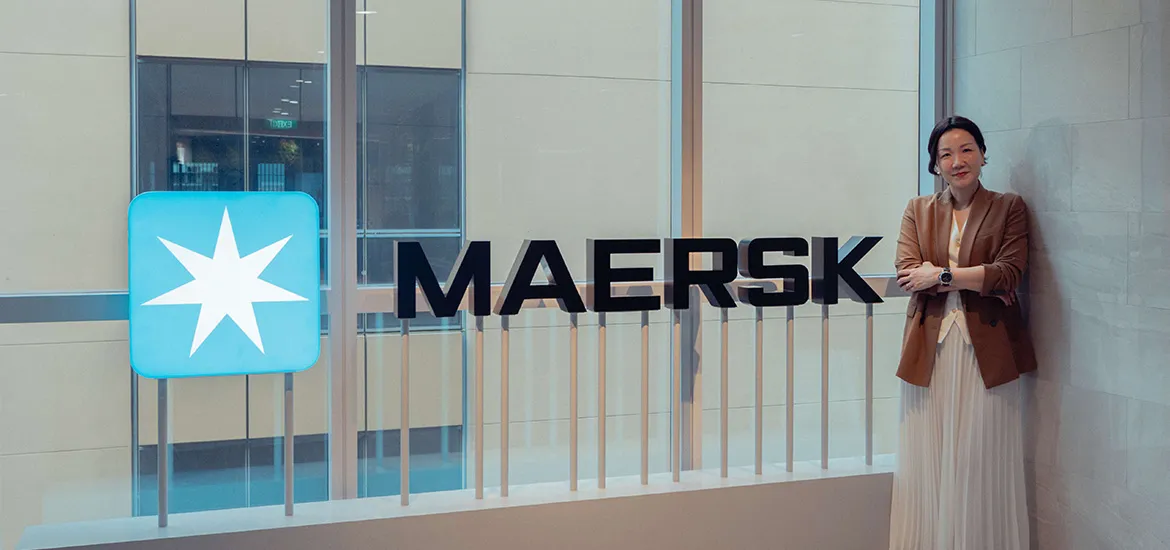 A woman stands beside a large illuminated Maersk logo inside a modern office with floor-to-ceiling windows.