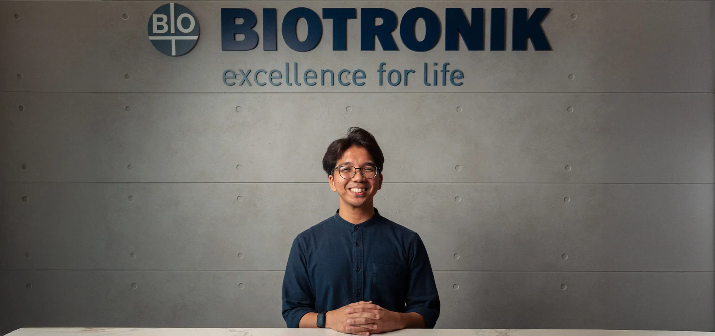 A smiling man in a dark shirt and glasses sits at a reception desk with his hands folded. Behind him is a grey wall with the logo and slogan of BIOTRONIK, a medical technology company. The logo includes the word "BIO" in a circular emblem and the text reads "BIOTRONIK – excellence for life."