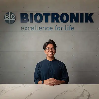 A smiling man in a dark shirt and glasses sits at a reception desk with his hands folded. Behind him is a grey wall with the logo and slogan of BIOTRONIK, a medical technology company. The logo includes the word "BIO" in a circular emblem and the text reads "BIOTRONIK – excellence for life."