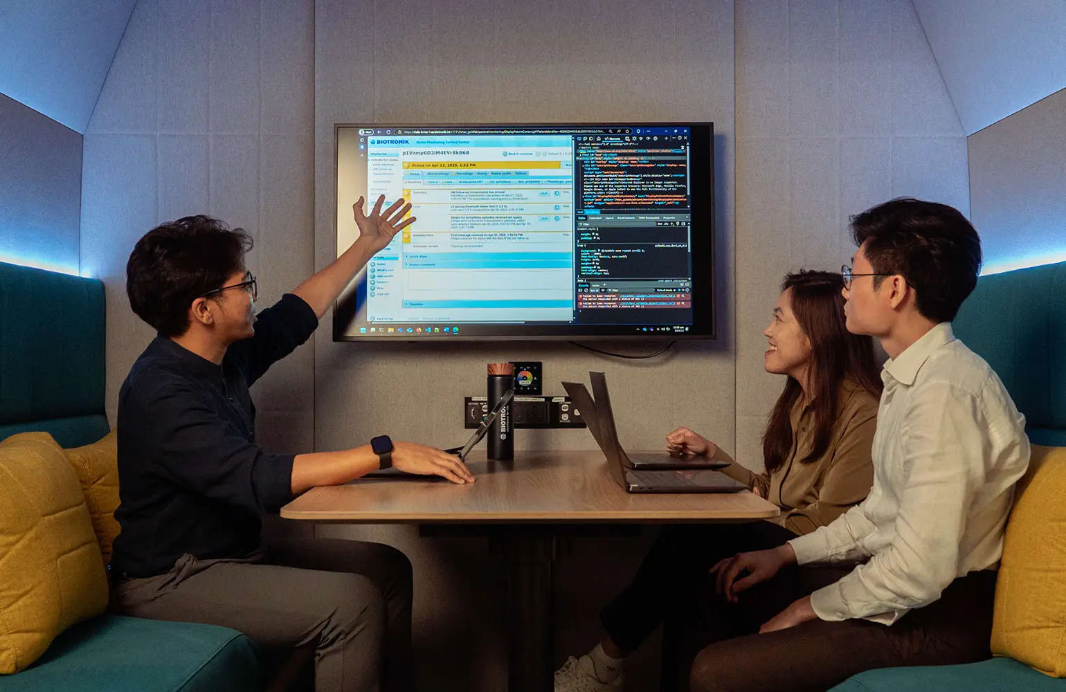 Three young professionals are in a modern meeting booth. One man on the left is presenting information displayed on a wall-mounted screen showing code and a web interface. A woman and another man are seated on the right, both engaged and smiling, with a laptop on the table. The setting is casual and collaborative, with cushioned walls and warm lighting.