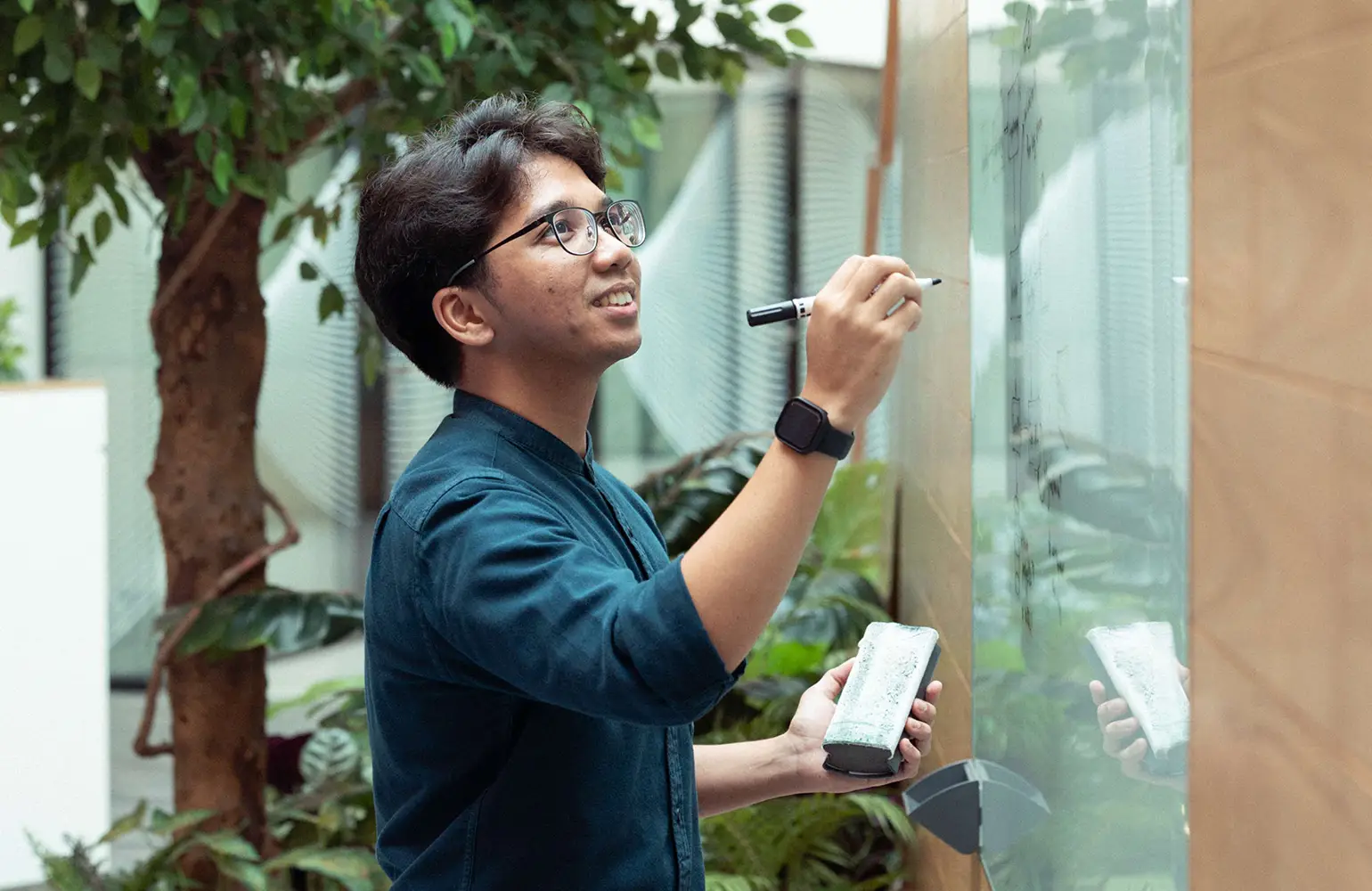 A young man with glasses is smiling while writing on a transparent whiteboard with a black marker. He holds an eraser in his other hand and wears a smart casual outfit with a smartwatch. The background includes indoor plants, creating a bright and modern office atmosphere.