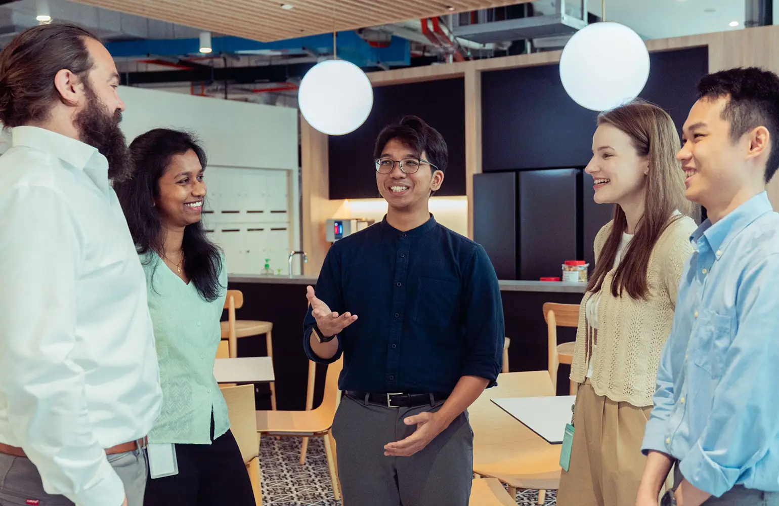 A diverse group of five professionals, including three men and two women, are standing in a modern office pantry area, engaged in a lively and friendly conversation. The central figure is smiling and gesturing animatedly while others are attentively listening and smiling. The setting includes contemporary lighting and furniture.