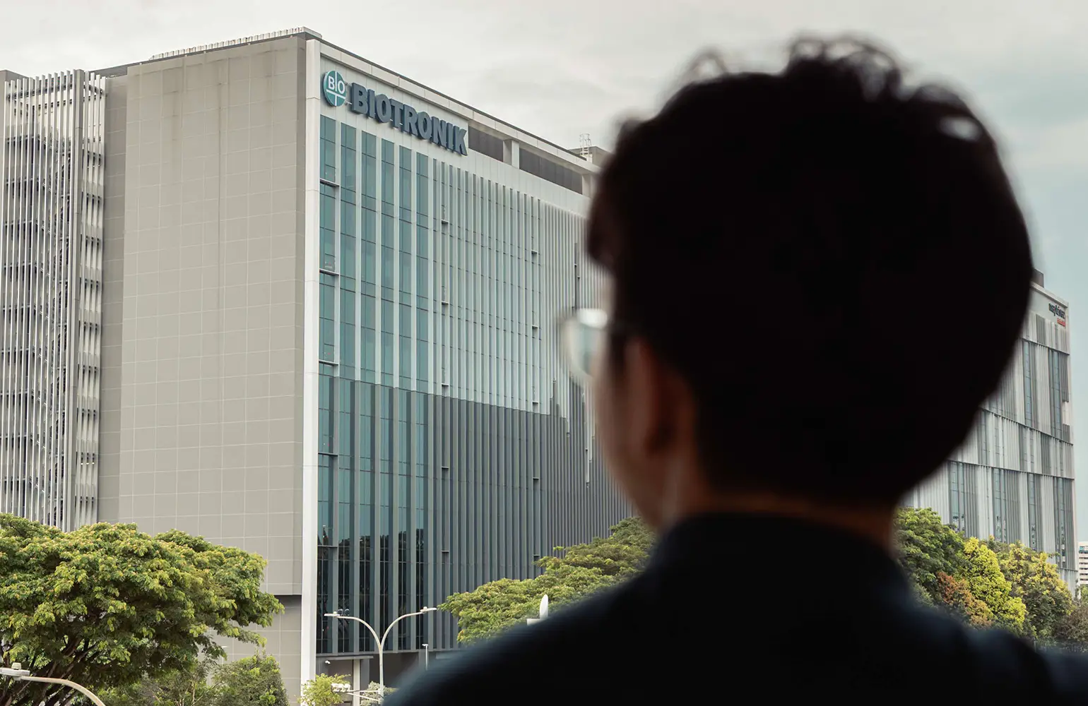 A person viewed from behind looks out at a modern glass-fronted building with the BIOTRONIK logo on top. The scene is set in an urban environment with trees and streetlights in the foreground, emphasizing a professional and corporate setting.