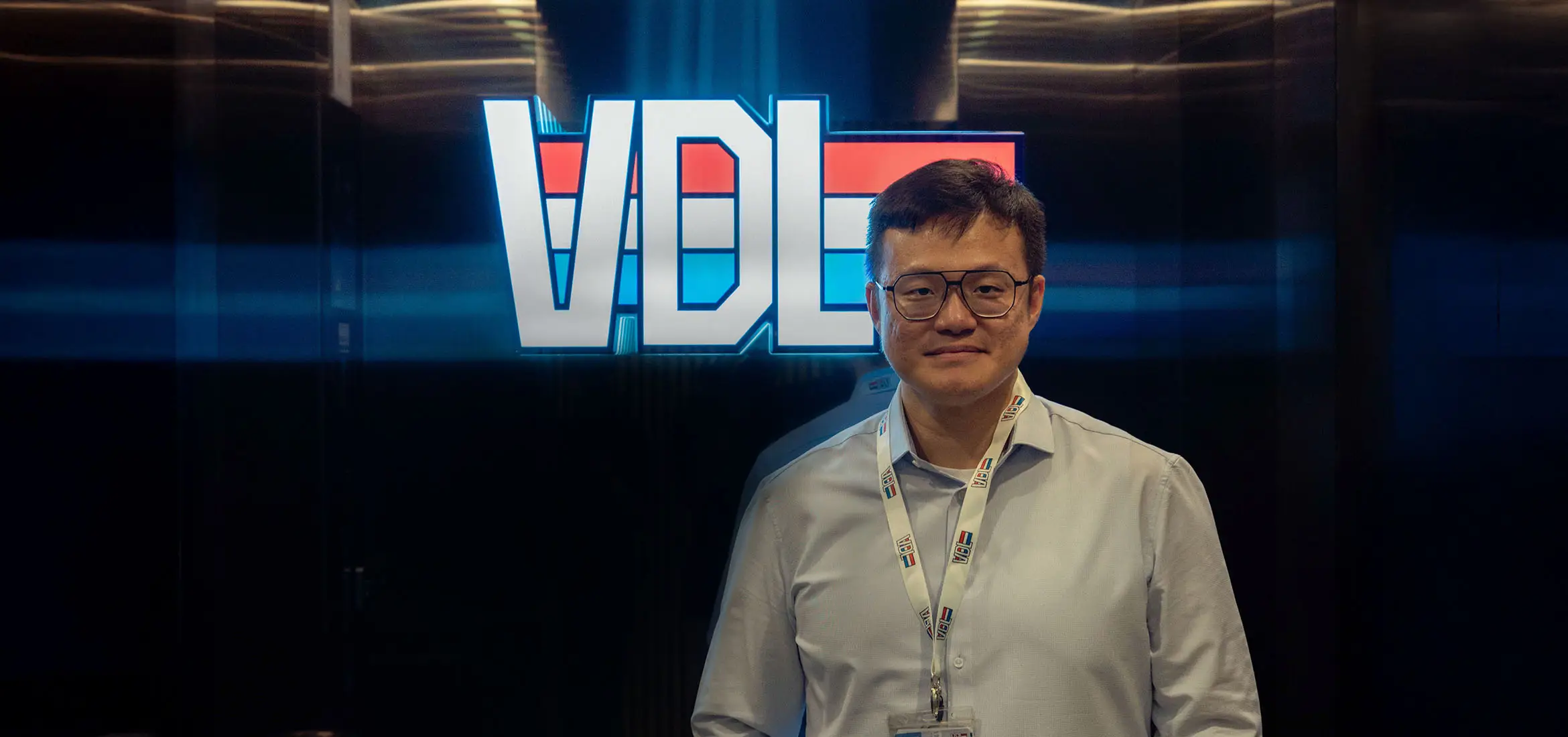 A man wearing glasses and a white shirt stands confidently in front of a brightly illuminated "VDL" logo. He is wearing an ID badge around his neck, suggesting a professional or corporate setting.