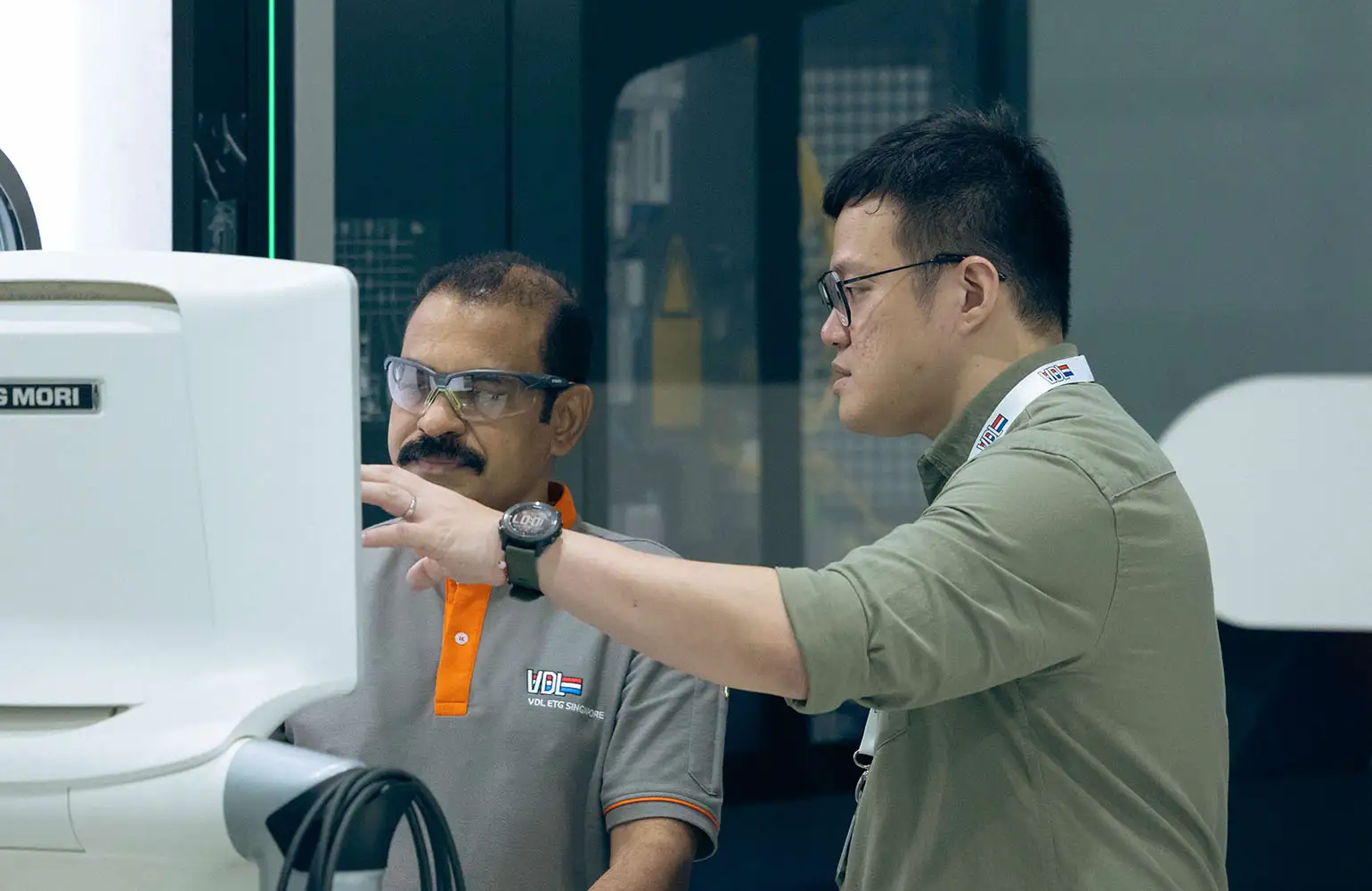 Two VDL engineers collaborating at a workstation. One points at a screen while the other listens attentively, both wearing safety glasses and VDL uniforms in a high-tech industrial setting.
