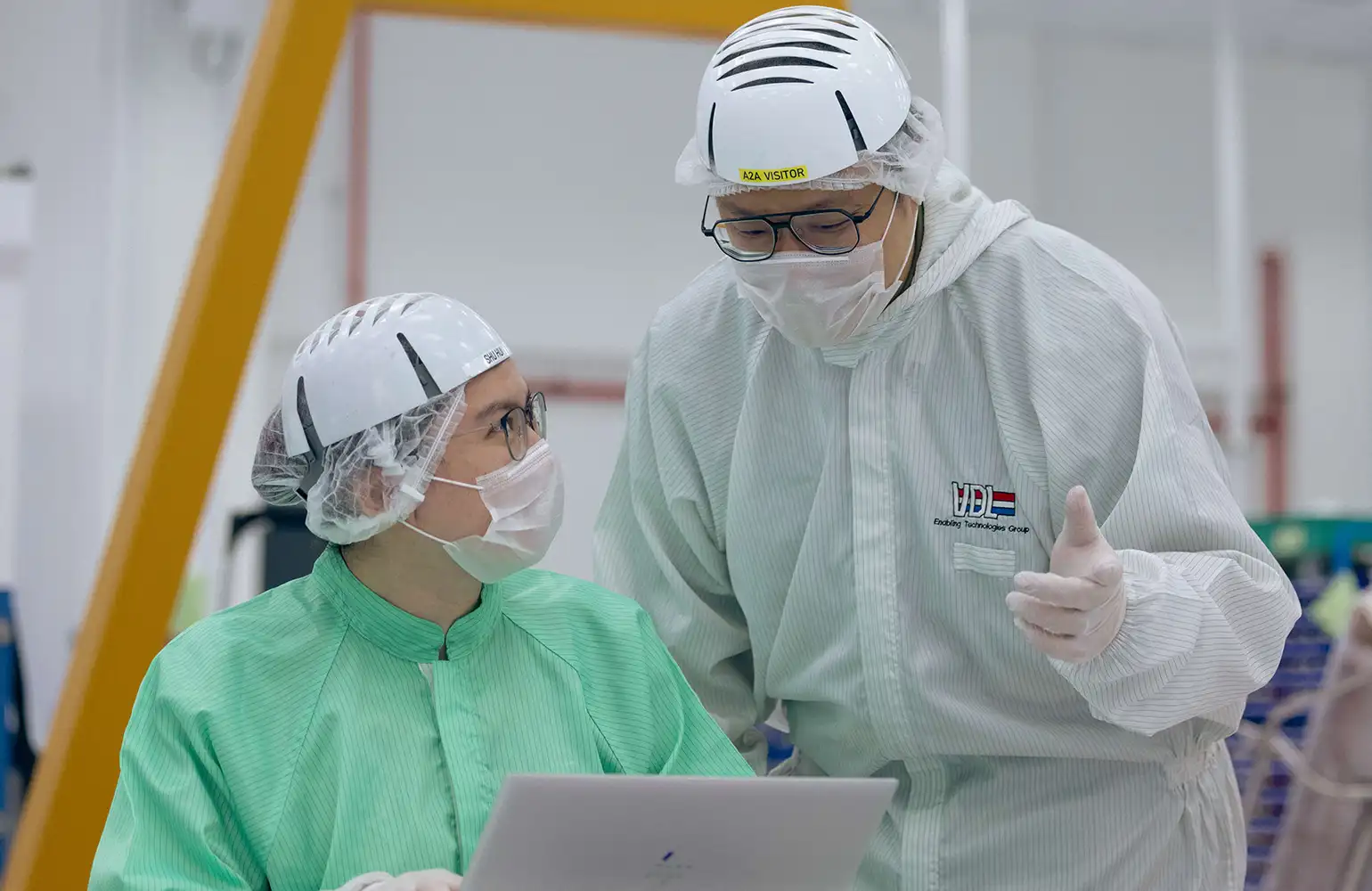 Two employees in cleanroom suits and safety helmets engage in a technical discussion while reviewing data on a laptop. The individual standing gestures while speaking, as the seated colleague attentively looks on, indicating a collaborative work environment in a high-tech manufacturing facility.