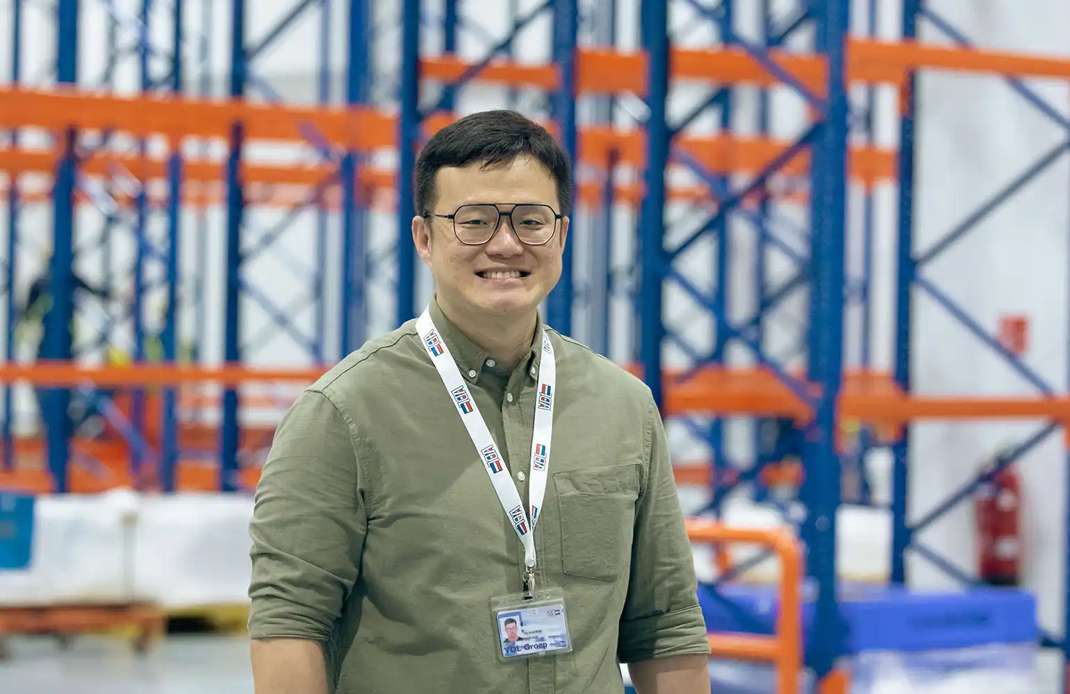 A smiling man in glasses and a green shirt stands confidently in front of industrial warehouse shelving. He wears a lanyard with an ID badge, suggesting his professional role within the facility. The setting conveys a clean, organized, and modern work environment.