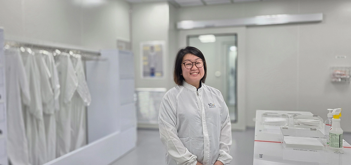 A technician in a cleanroom suit stands smiling inside a laboratory, with equipment and protective garments visible in the background.