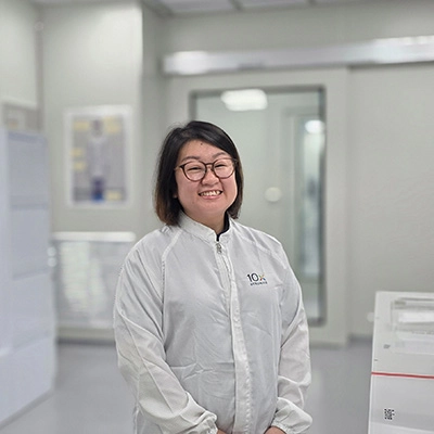A technician in a cleanroom suit stands smiling inside a laboratory, with equipment and protective garments visible in the background.