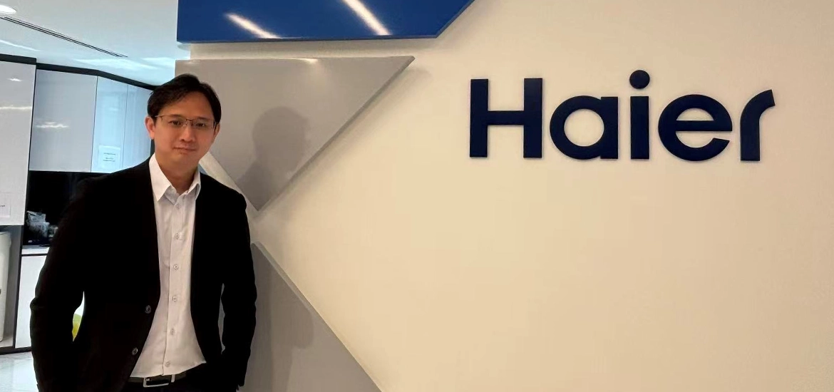 A man in business attire standing beside a wall with the Haier company logo inside a modern office setting.