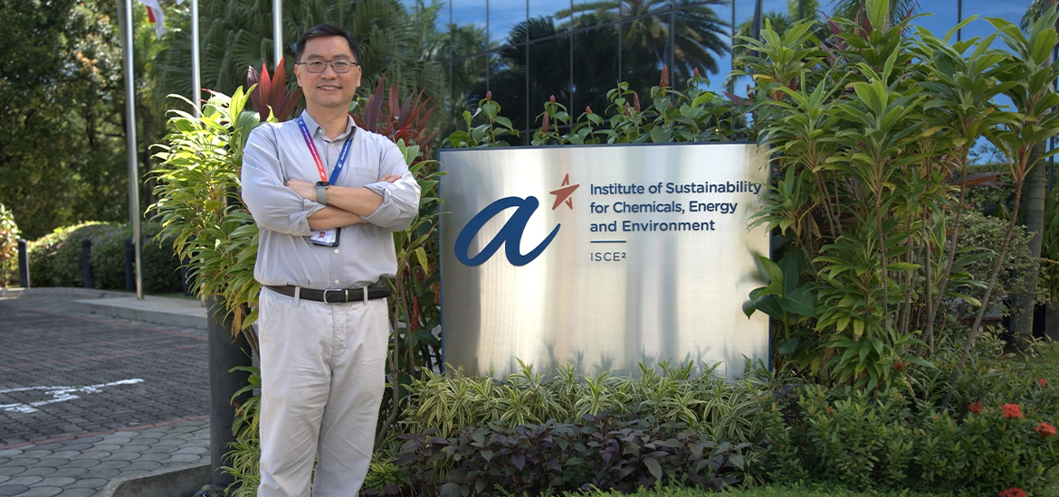 Man standing with folded arms beside the signboard of ASTAR’s Institute of Sustainability for Chemicals, Energy and Environment (ISCE²), surrounded by greenery outside the facility.*