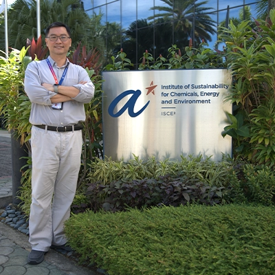 Man standing with folded arms beside the signboard of ASTAR’s Institute of Sustainability for Chemicals, Energy and Environment (ISCE²), surrounded by greenery outside the facility.*