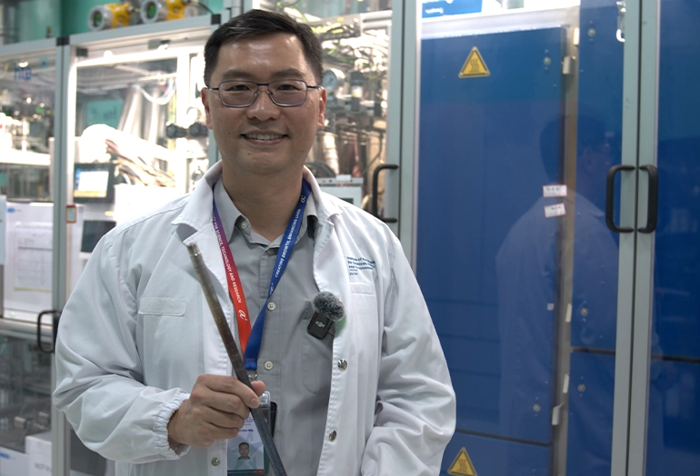 Scientist wearing a lab coat and ID badge smiles while holding a metallic sample inside a laboratory with industrial equipment in the background.