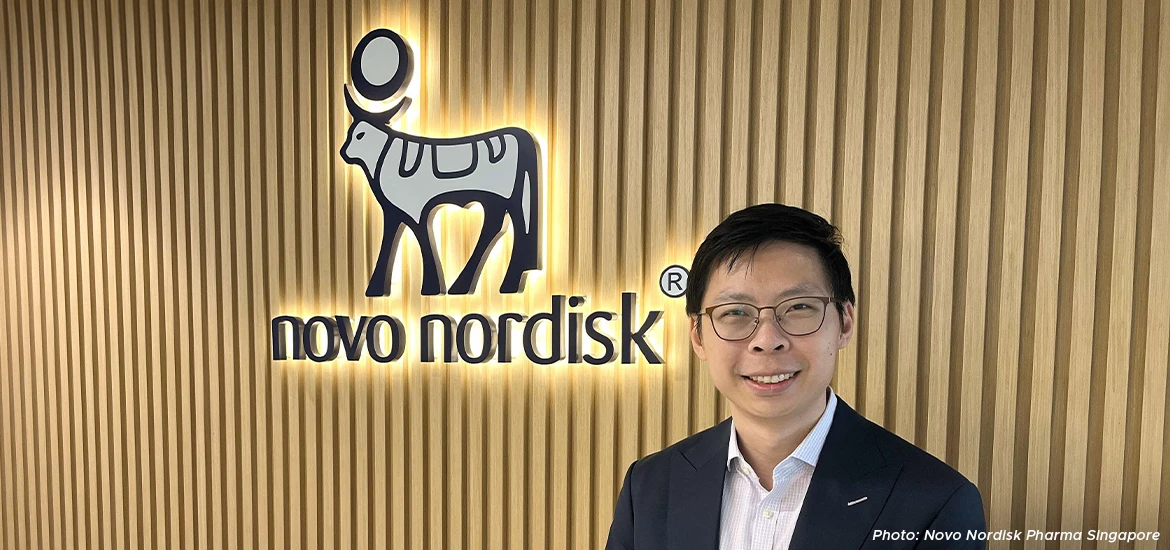 Smiling professional standing in front of the Novo Nordisk logo wall, symbolizing leadership and innovation in the healthcare and pharmaceutical industry.