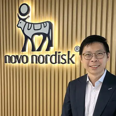 Smiling professional standing in front of the Novo Nordisk logo wall, symbolizing leadership and innovation in the healthcare and pharmaceutical industry.