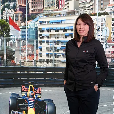 A woman wearing a black Mobil shirt stands outdoors, smiling, with city buildings and a red-and-white flag in the background.