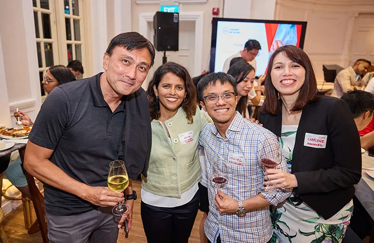 Four people smiling and posing together at a networking event, holding drinks in a brightly lit indoor venue.