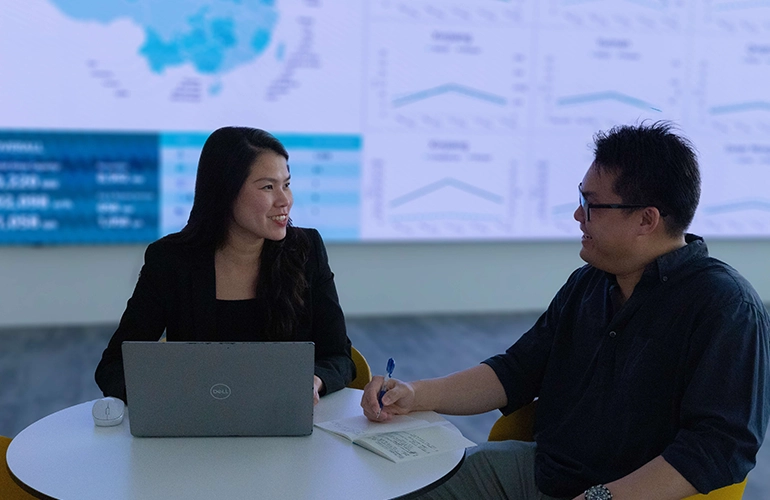 Two professionals discuss data at a table, with charts and analytics displayed on a screen behind them.