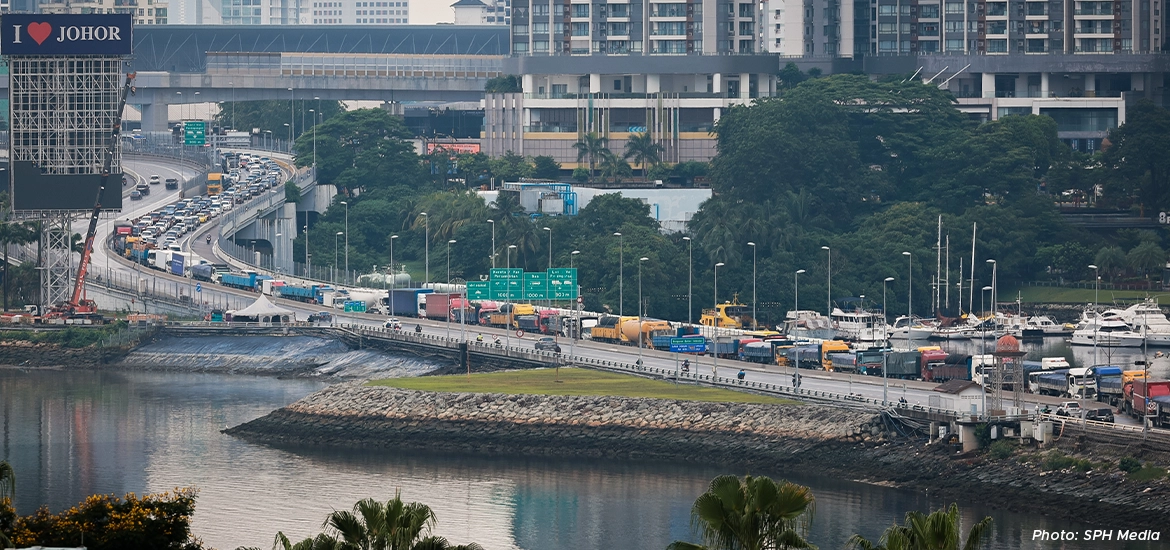 : A busy causeway connecting Singapore and Johor filled with cars and trucks, with city buildings and greenery in the background.