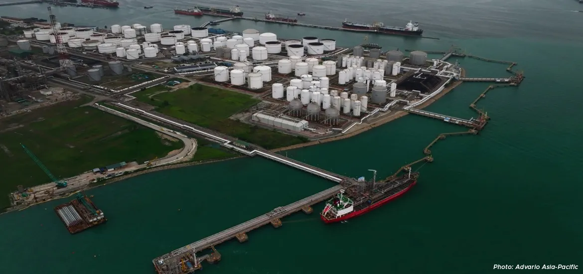 Aerial view of an industrial oil and chemical storage terminal with numerous white storage tanks, pipelines, and docked cargo ships along the waterfront.