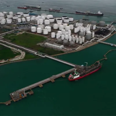 Aerial view of an industrial oil and chemical storage terminal with numerous white storage tanks, pipelines, and docked cargo ships along the waterfront.