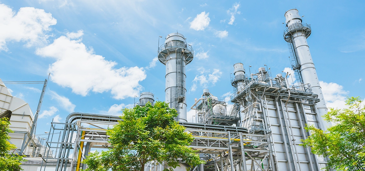 Modern power generation facility with tall gas turbine stacks, steel piping, and industrial structures set against a bright blue sky, with greenery in the foreground highlighting sustainable energy infrastructure.